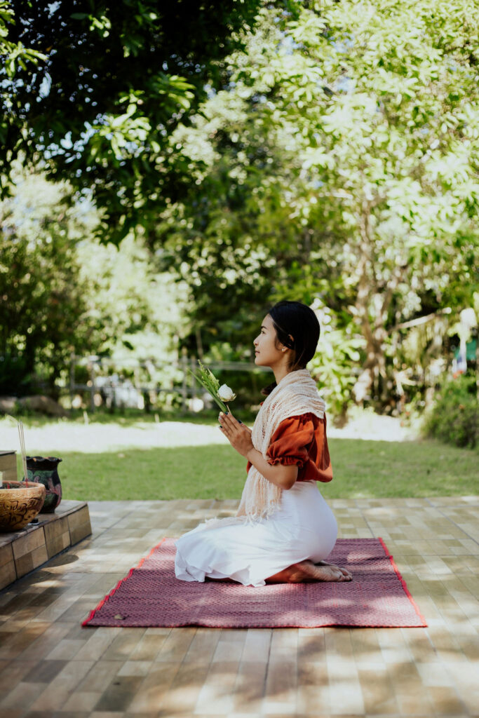 Woman kneeling on a mat outdoors, holding flowers in a peaceful prayer position